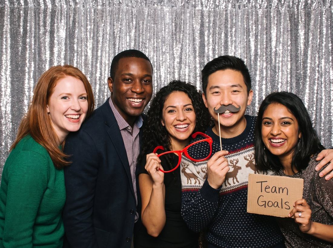 Five coworkers laughing with props in front of a silver sequin backdrop